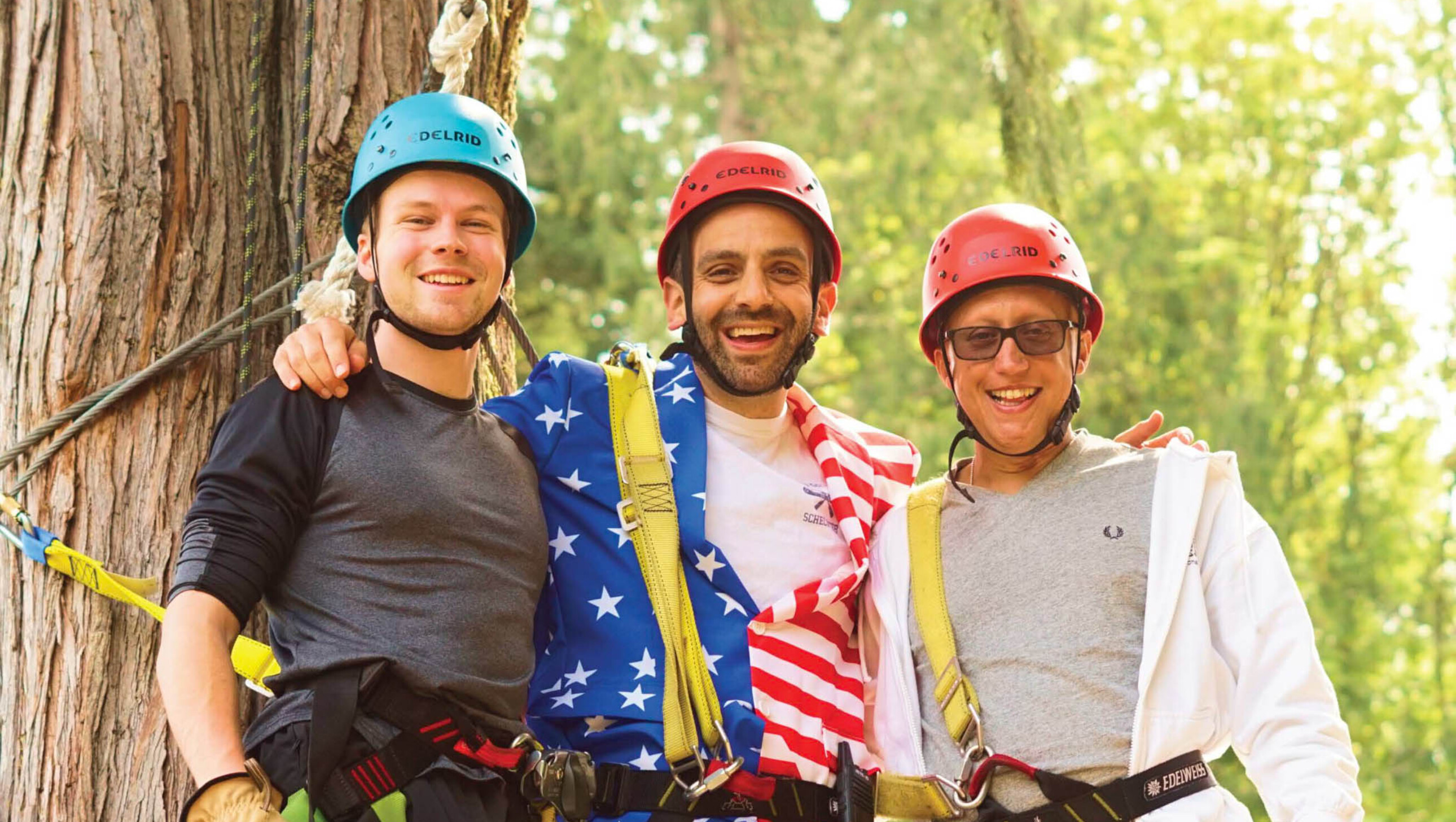 three men in harnesses and helmets next to a tree.